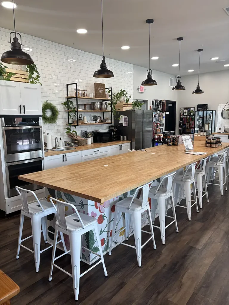 A bright, modern teaching kitchen inside Toast & Table, featuring a large wooden island with white floral-patterned base, surrounded by white metal stools. The backdrop includes white subway tile walls, open shelving with cookware and plants, and hanging industrial-style pendant lights. Retail shelves with kitchen goods are visible in the background.