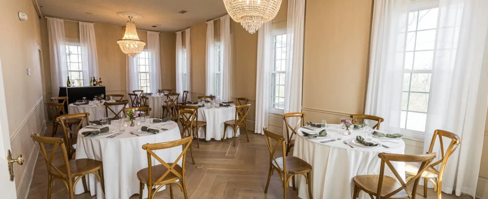 Cozy dining room with wooden tables and chairs set with minimalist place settings and glassware, featuring hardwood floors and large windows.