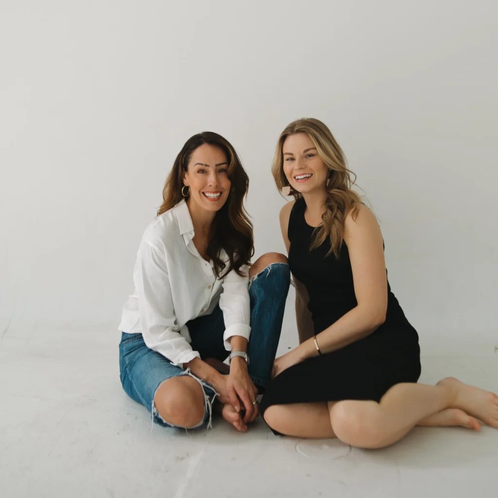 Brandi Radny and Brittany Nash sit on the floor in a minimalist studio with a white background.