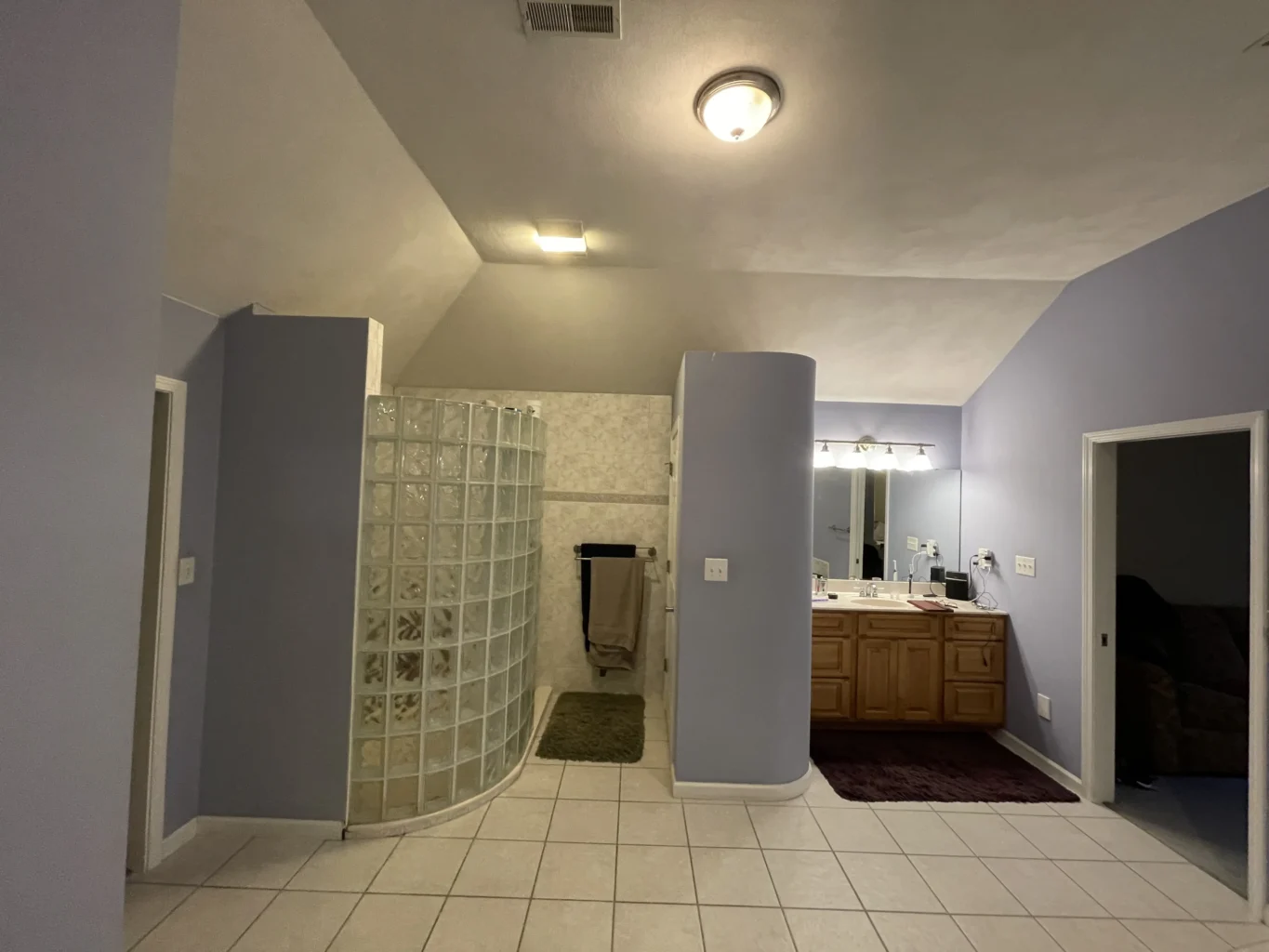 Before of an outdated bathroom with a curved glass block shower, white tile flooring, vanity and dim lighting.
