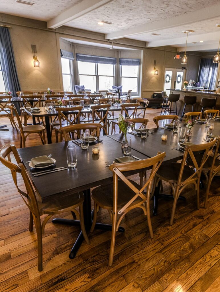 Cozy dining room with wooden tables and chairs set with minimalist place settings and glassware, featuring hardwood floors and large windows.